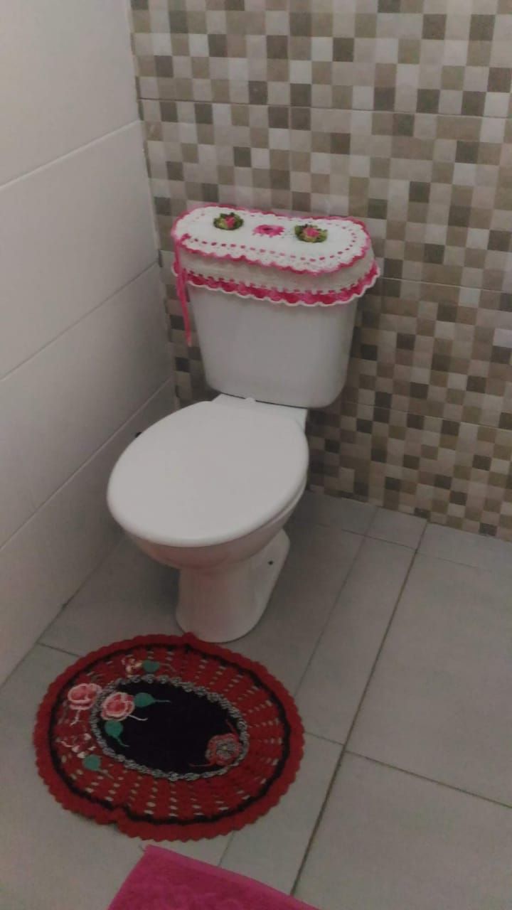 Des toilettes dotées d’un couvercle décoratif rose et blanc, un mur carrelé avec un motif à carreaux et un tapis rouge en crochet sur le sol.