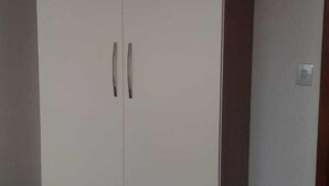 Ein großer, weißer Kleiderschrank mit zwei Türen und einem hölzernen Sockel, vor einer schlichten Wand in einem Raum mit Fliesenboden platziert.