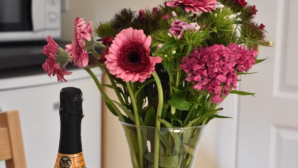 Eine Prosecco-Flasche, eine Packung mit verklärten Fingerkeks, ein Schlüsselanhänger und eine Vase mit rosa Blumen auf einem Tisch.