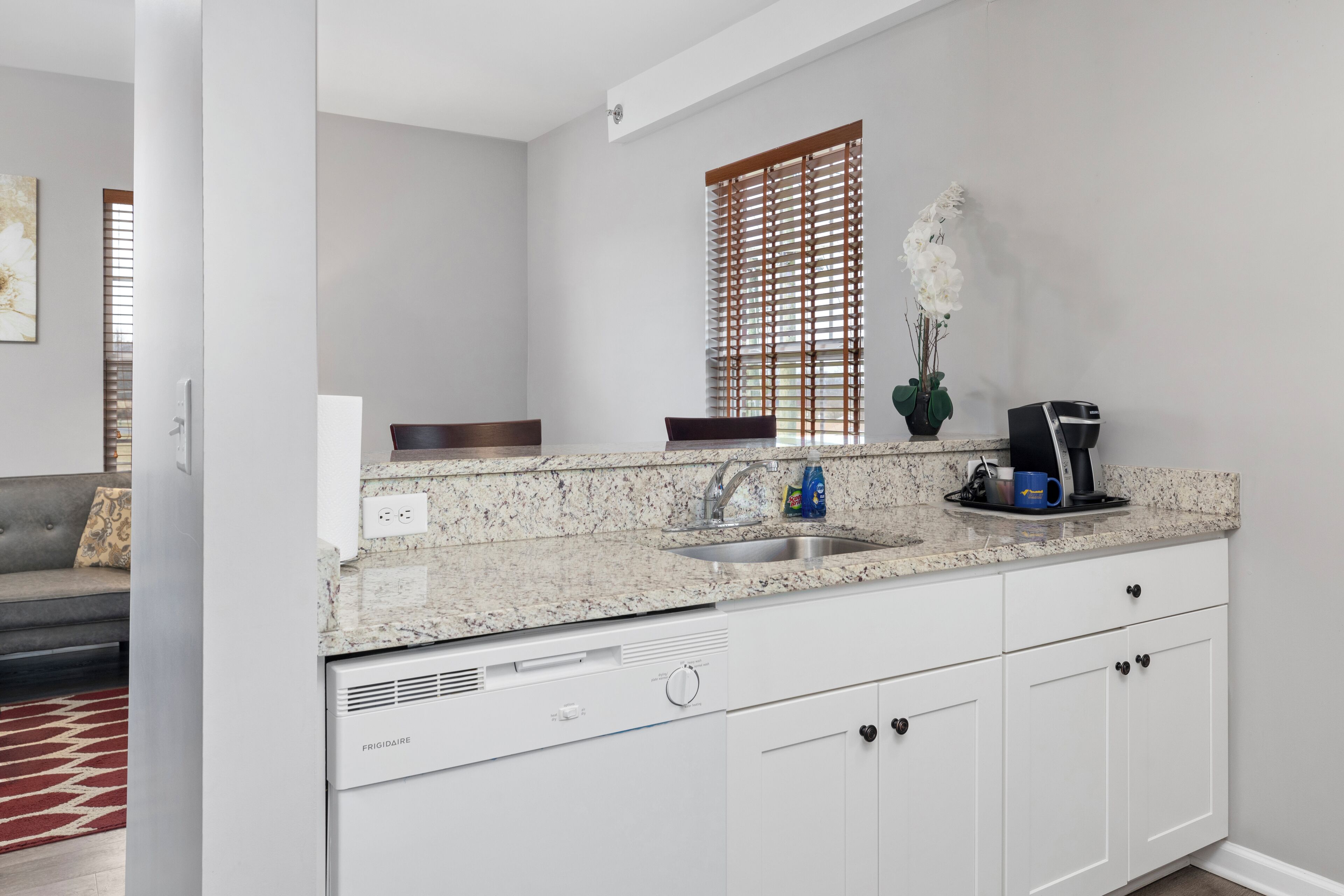 A modern kitchen with white cabinetry, a granite countertop, a built-in dishwasher, and a window with blinds.