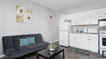 Una sala de estar moderna con un sofá gris, una mesa de centro negra y una cocina blanca con armarios y electrodomésticos blancos.