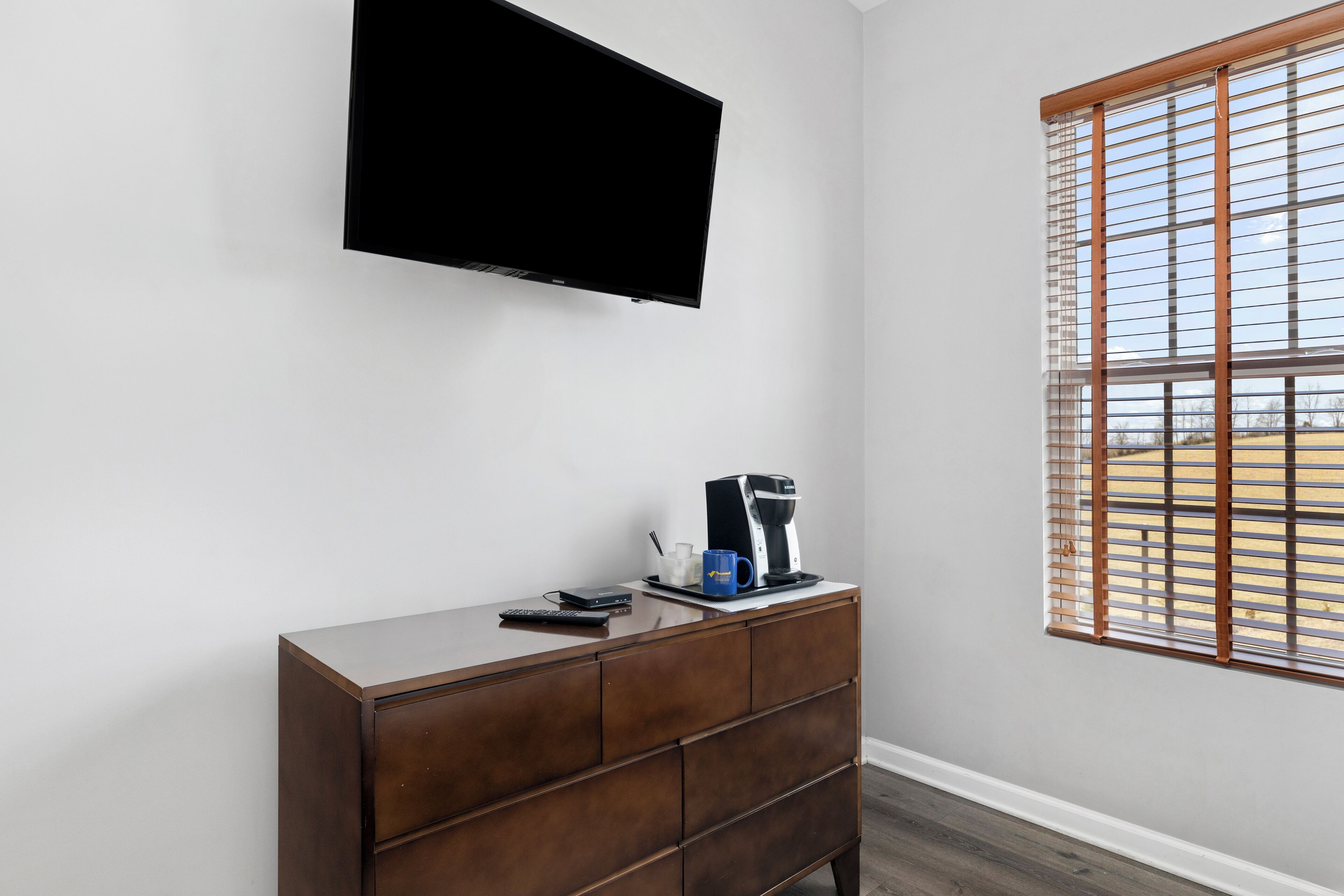 Una habitación con una televisión de pantalla plana en la pared, un aparador de madera con cajones, una cafetera y una ventana con persianas.