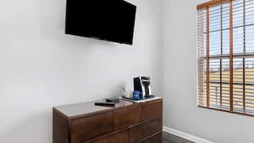 Una habitación con una televisión de pantalla plana en la pared, un aparador de madera con cajones, una cafetera y una ventana con persianas.