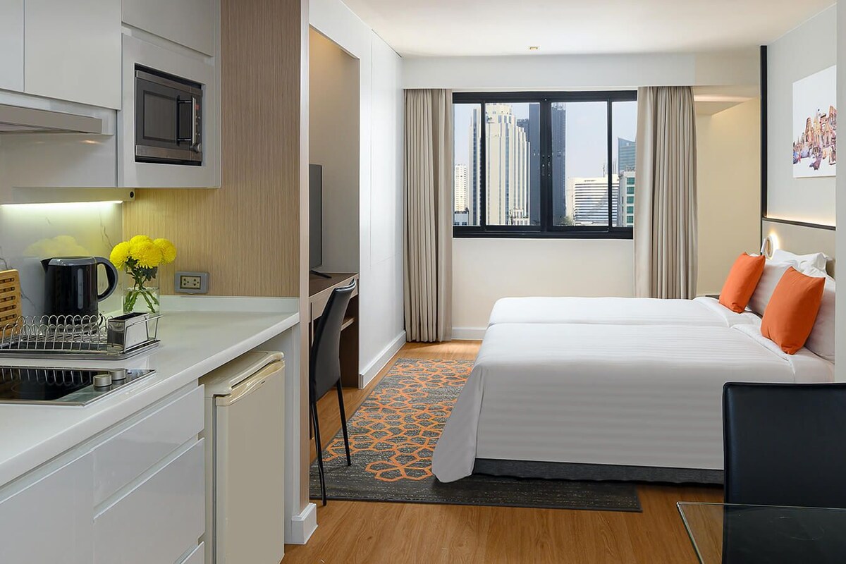 Habitación de hotel con kitchenette, una cama con almohadas naranjas, un escritorio con silla y ventana con vista a la ciudad.