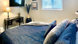 A bedroom with a bed, a TV mounted on the wall, a window with blinds, a bedside table with a lamp, and a decorative vase. - Beautifully remodeled, clean, well-stocked entire home - sleeps 7! (Yakima)