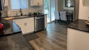 A modern kitchen with white cabinets, a black countertop, and a built-in dishwasher. There is a dining area with a table and chairs, and a large window with blinds. - Beautifully remodeled, clean, well-stocked entire home - sleeps 7! (Yakima)