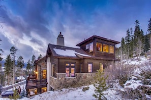 A large, multi-story wooden house with stone accents, surrounded by snow and trees.