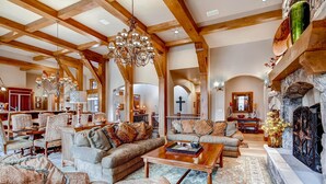 Ein geräumiges Wohnzimmer mit hohen Decken, Holzbalken und einem Chandelier. Ein Kamin mit Dekorationselementen und eine Steinwand sind ebenfalls vorhanden.