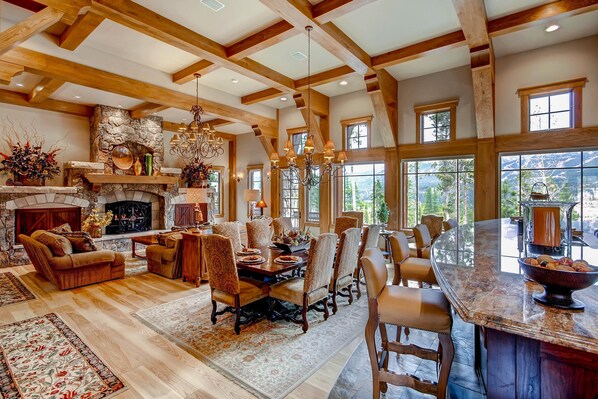 A spacious living room with a fireplace, wooden beams, large windows, and a dining area with a marble island.
