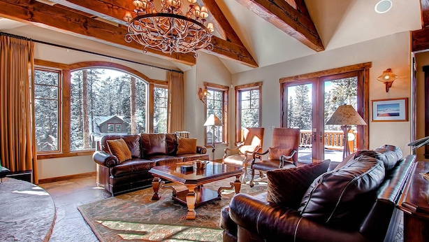 A spacious living room with a large wooden ceiling, leather sofas, a wooden coffee table, and large windows offering a view of the forest. - Rustic Timber Lodge (Breckenridge)