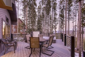 A wooden deck with outdoor furniture, surrounded by tall trees and a view of a mountainous landscape.