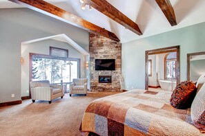 A spacious bedroom with a large bed, a stone fireplace, a television, and a view of the outdoors.