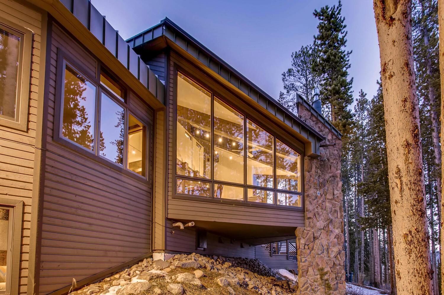 A modern house with large windows, stone foundation, and a view of a forest.