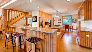 A spacious kitchen with wooden cabinets, a central island with stools, and a staircase with wooden railings. - Inglenook (Breckenridge)