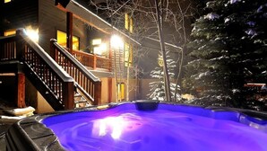 A hot tub in a snowy outdoor setting with a wooden deck and a building in the background. - Inglenook (Breckenridge)