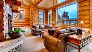 L’intérieur d’une cabane en rondins, avec une cheminée en pierre, des canapés en cuir et de grandes fenêtres offrant une vue panoramique.