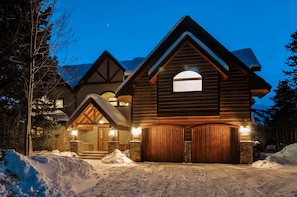 A large log cabin with a steeply pitched roof, a central chimney, and a stone foundation.