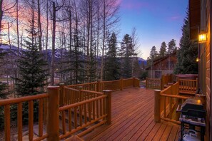 A wooden deck with a railing, overlooking a forested area with tall trees and a mountain in the background.
