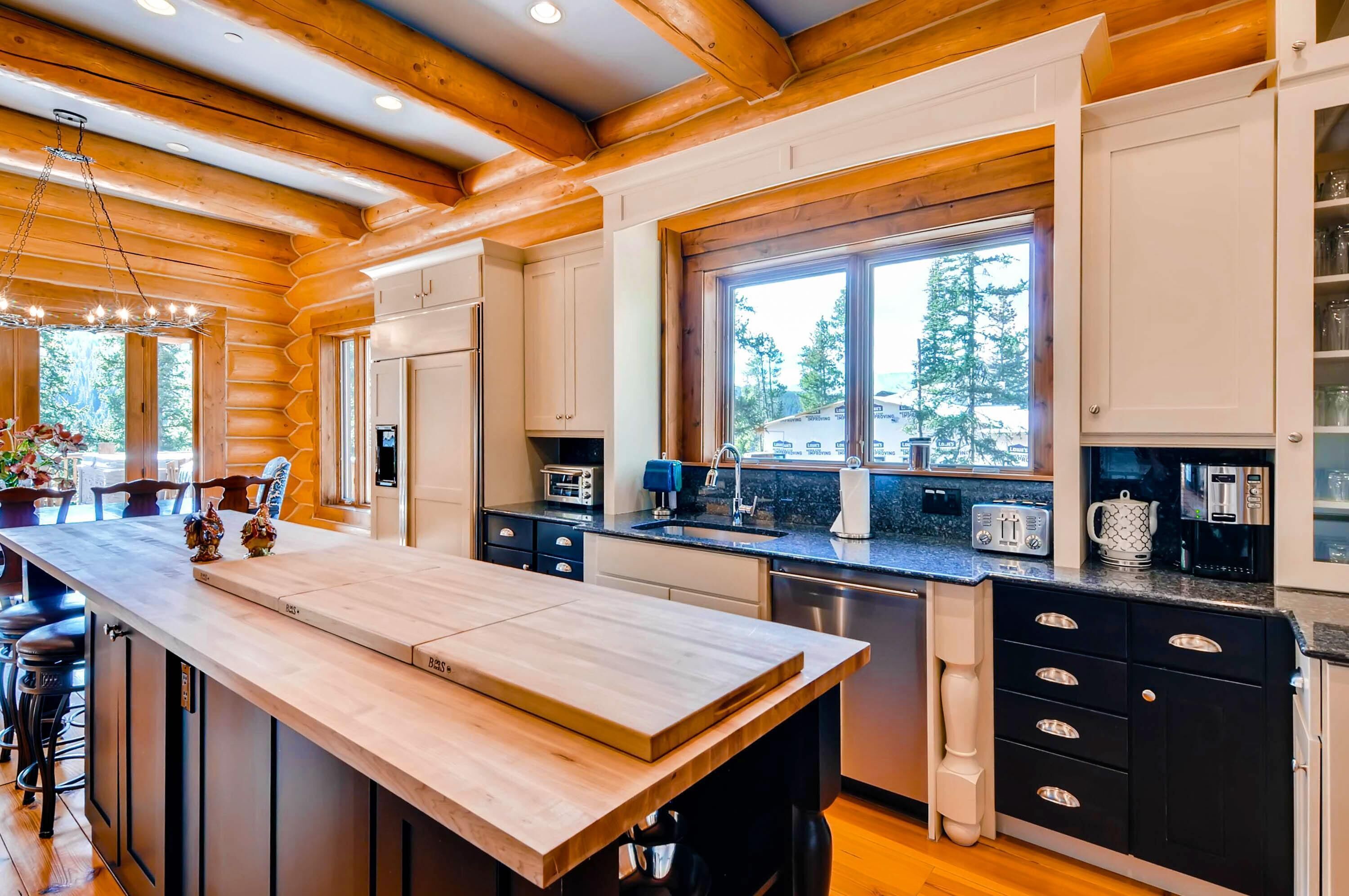 A modern kitchen with wooden beams, a large island, and a window overlooking a forest.