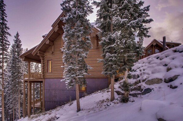 A log cabin with a balcony, surrounded by snow-covered trees and a rocky hillside.