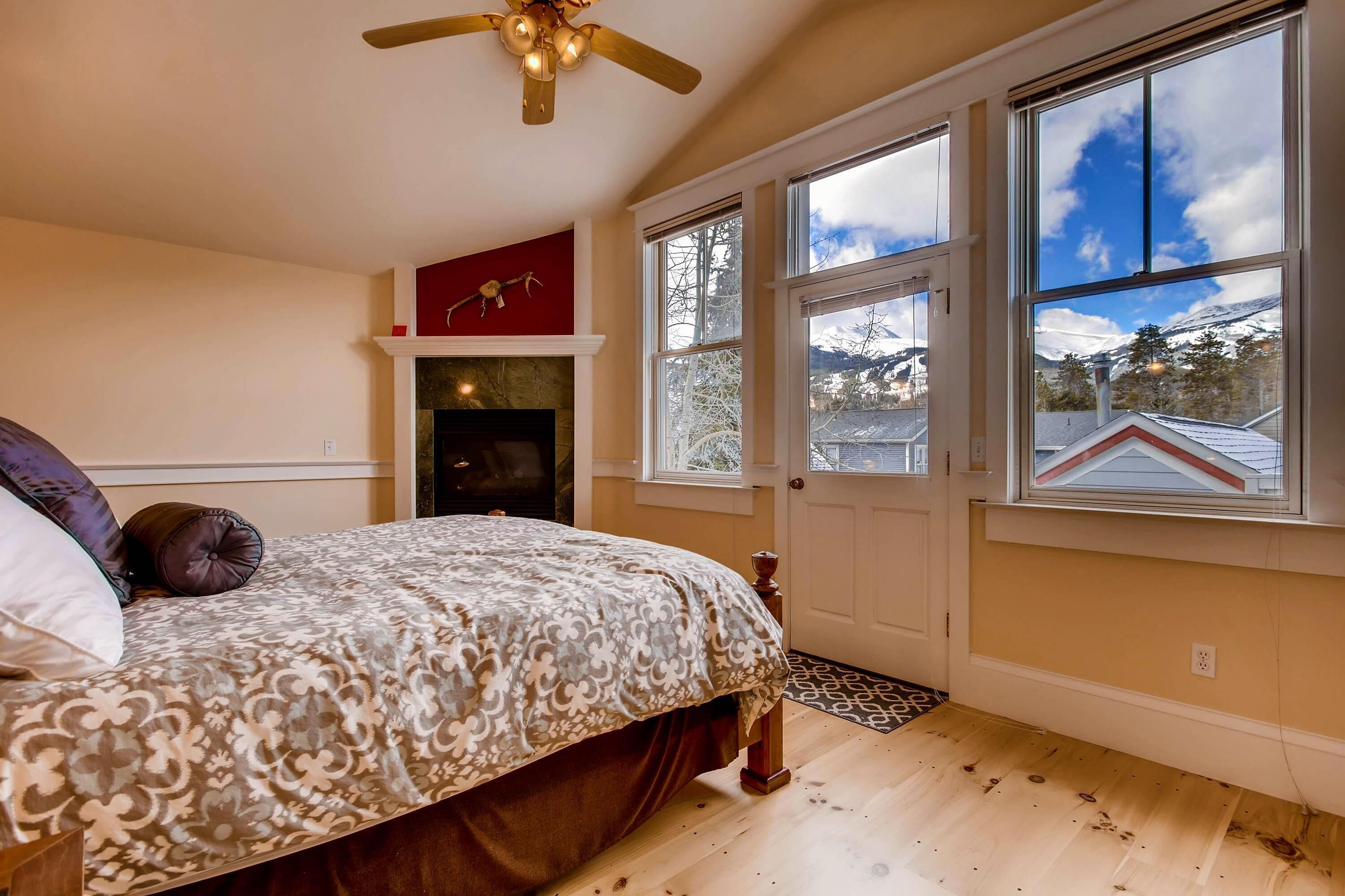 A bedroom with a large bed, a fireplace, and a view of the outdoors through large windows.
