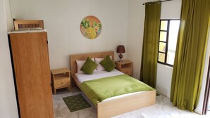 Ein Schlafzimmer mit einem hölzernen Bett, grünen Kissen, einem Nachttisch mit Lampe, einem kleinen Tisch und einem Fenster mit grünen Vorhängen.