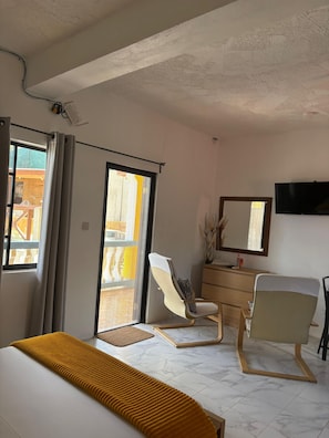 Une chambre d’hôtel comprenant un lit, deux chaises, une commode, un téléviseur et une porte-fenêtre coulissante donnant sur un balcon.