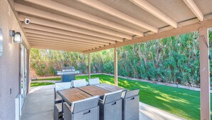 An outdoor patio with a wooden table and wicker chairs, a barbecue grill, and a view of a well-maintained lawn and trees. - Welcome to your Dream Home, up to 12 guests! (Henderson)
