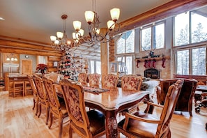 Une grande table à manger en bois, entourée de chaises richement décorées, d’un lustre et d’une cheminée agrémentée d’une console.