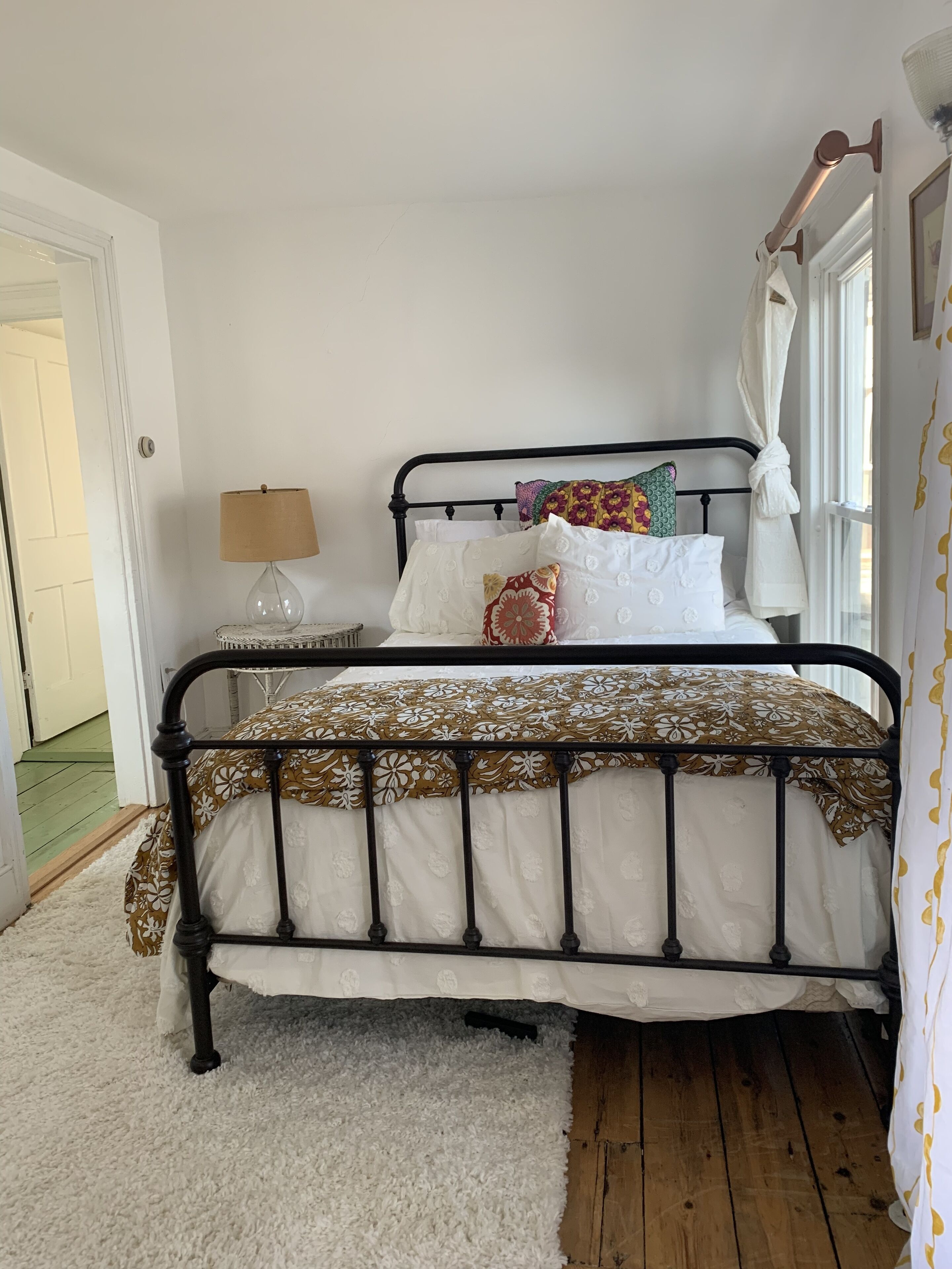 A bedroom with a metal bed frame, white bedding, a fluffy white rug, and a bedside table with a lamp.