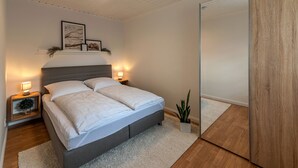 A bedroom with a bed, bedside tables, a nightstand, a wall-mounted shelf with framed pictures, a potted plant, and a mirror. - Ferienapartment Weber (Faßberg)