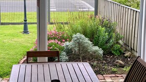 Um deck de madeira com mesa e cadeiras, com vista para um jardim com diversas plantas e um lago com barcos.