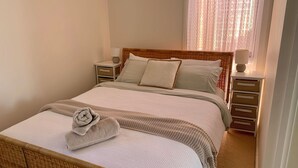 Cama bem arrumada com edredom branco e toalha decorativa. Há dois travesseiros e cabeceira com padrão trançado. Mesa de cabeceira com abajur e gavetinha visível.