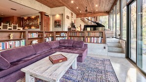 Uma sala de estar ampla com um sofá seccional roxo grande, uma mesa de centro de madeira e uma estante cheia de livros. Um piano de cauda é visível ao fundo, e o ambiente apresenta um teto alto com vigas de madeira e janelas grandes.