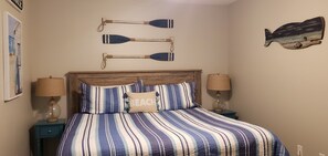 A bedroom with a striped bedspread, two bedside lamps, a wooden headboard, and wall art including paddle boats and a fish.