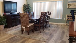 Un comedor con una mesa y sillas de madera, un televisor sobre un soporte, una planta grande en maceta y una ventana con persianas.