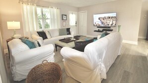 A living room with white furniture, a TV, and a window with curtains.