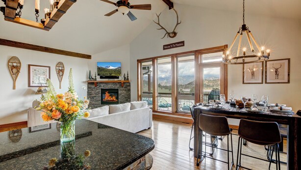 Un vaste espace de vie doté d’une cheminée, d’une table à manger avec des chaises, d’une grande fenêtre offrant une vue sur les montagnes et d’un ventilateur de plafond.