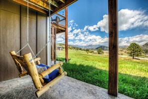 A wooden swing chair with a cushion, a balcony, and a view of a green field and mountains.