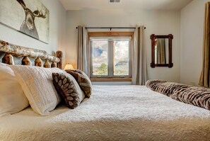 A bedroom with a large bed, a window with curtains, a wooden headboard, a mirror, and a wall-mounted picture.