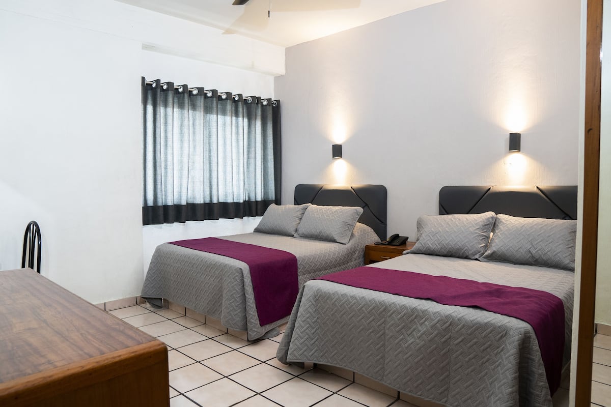 A hotel room with two beds, a wooden table, a window with curtains, and wall-mounted lights.