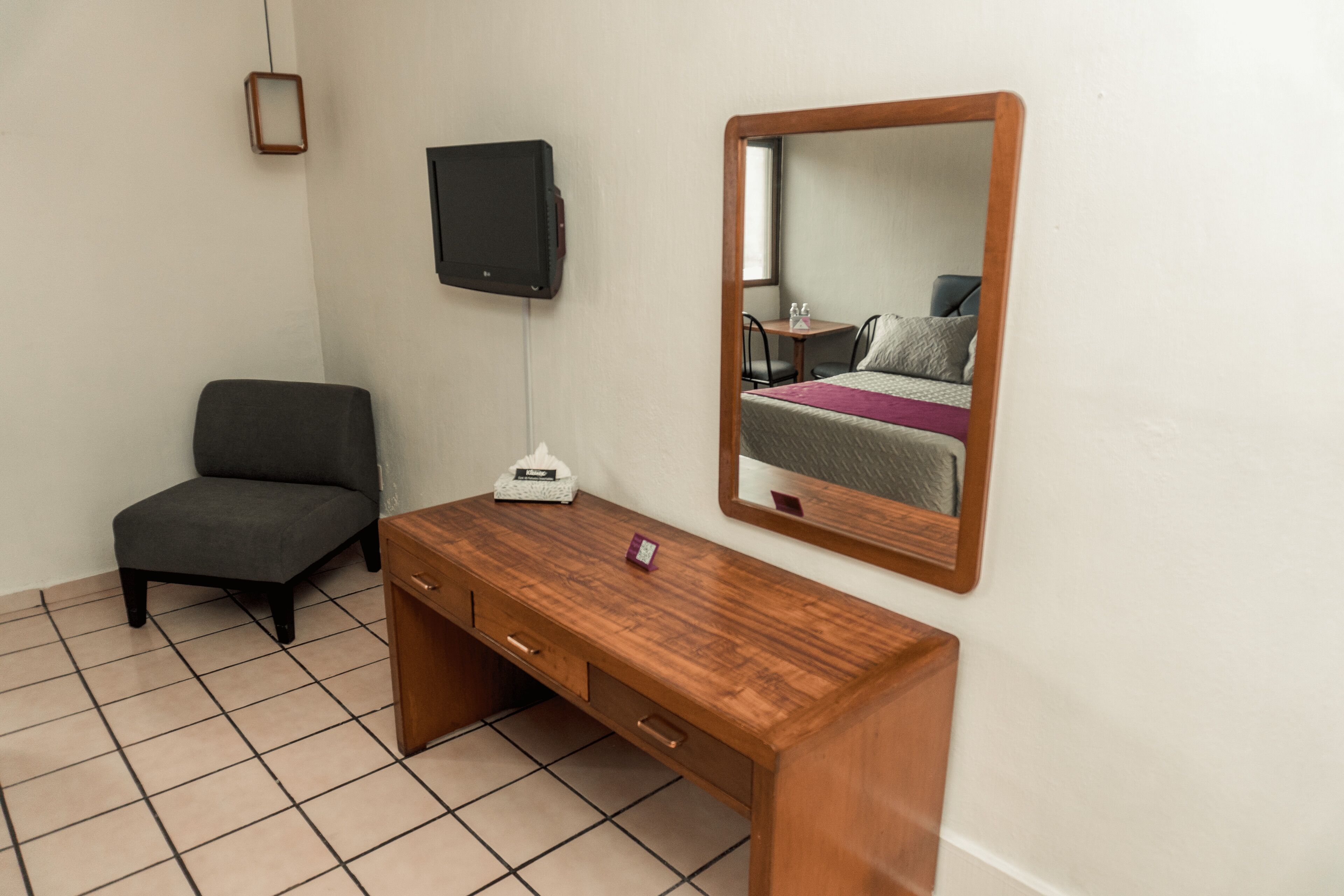 A small, tidy room with a wooden desk, a chair, a TV mounted on the wall, and a mirror reflecting the bed and window.