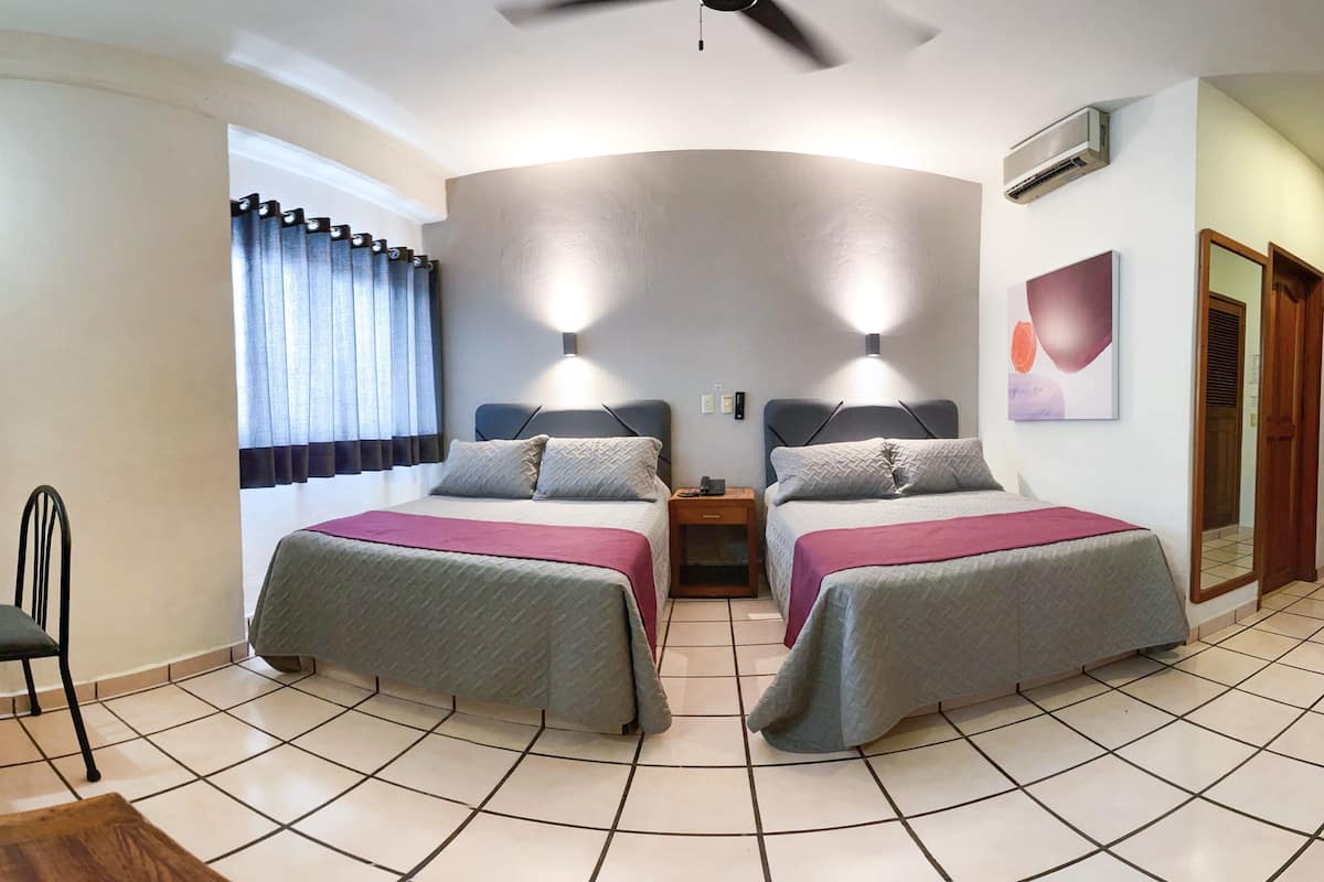 A room with two beds, a ceiling fan, a window with curtains, an air conditioner, and a painting on the wall.