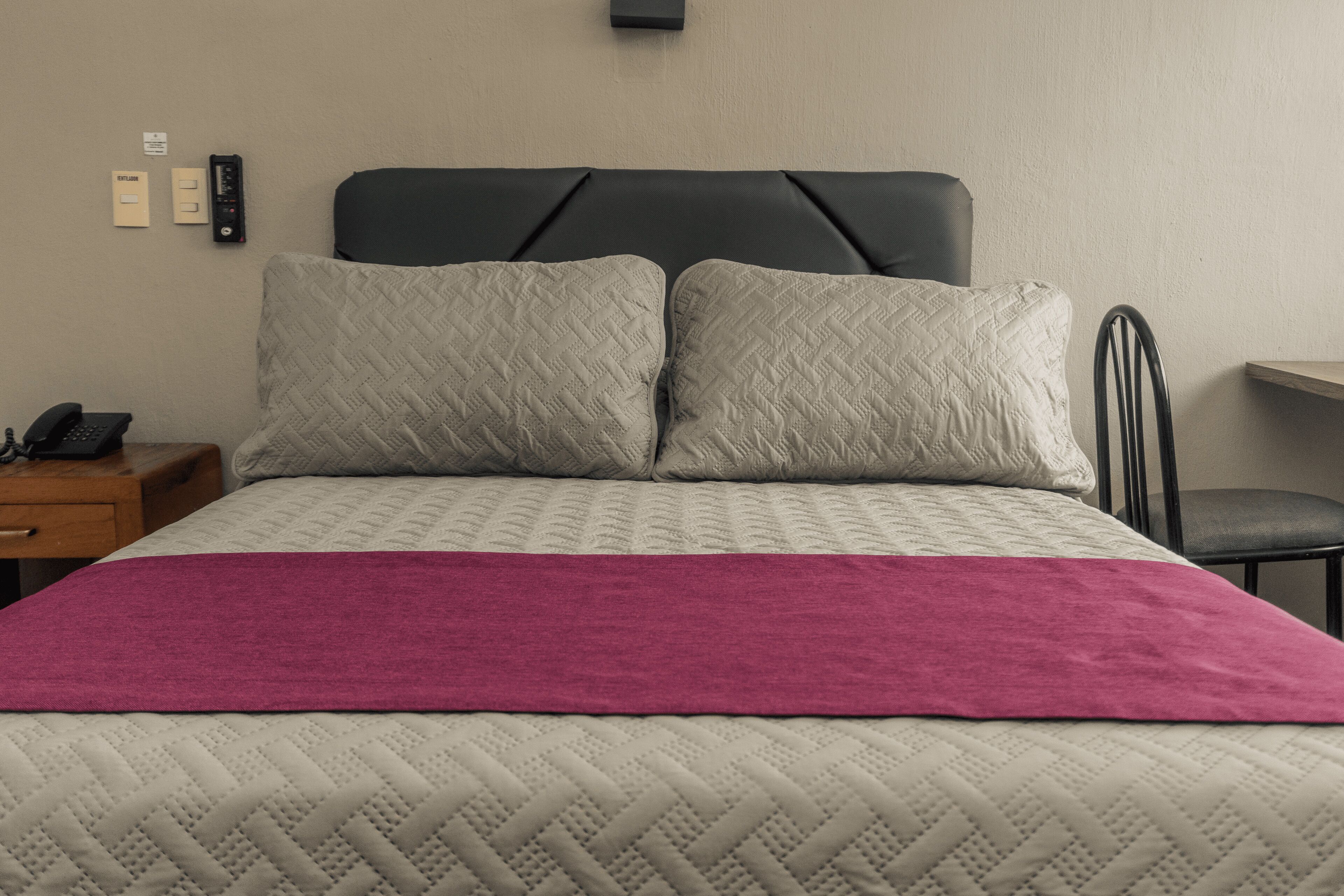 Een net opgemaakt bed met een gewatteerd hoofdbord, een roze deken en een nachtkastje met een telefoon.