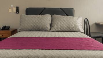 Een net opgemaakt bed met een gewatteerd hoofdbord, een roze deken en een nachtkastje met een telefoon.