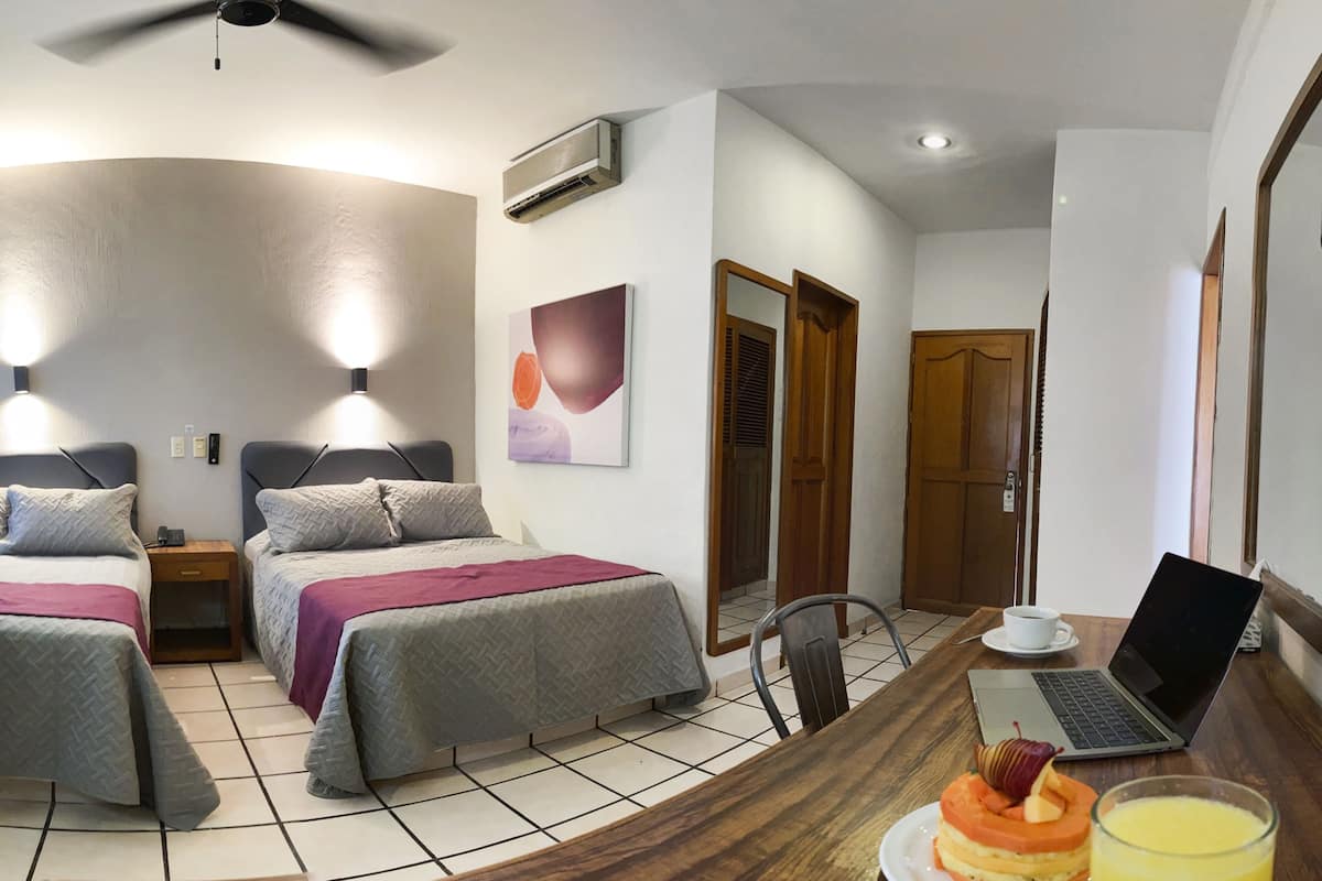 A hotel room with two beds, a desk with a laptop, a chair, a coffee cup, and a plate with food and a glass of juice.