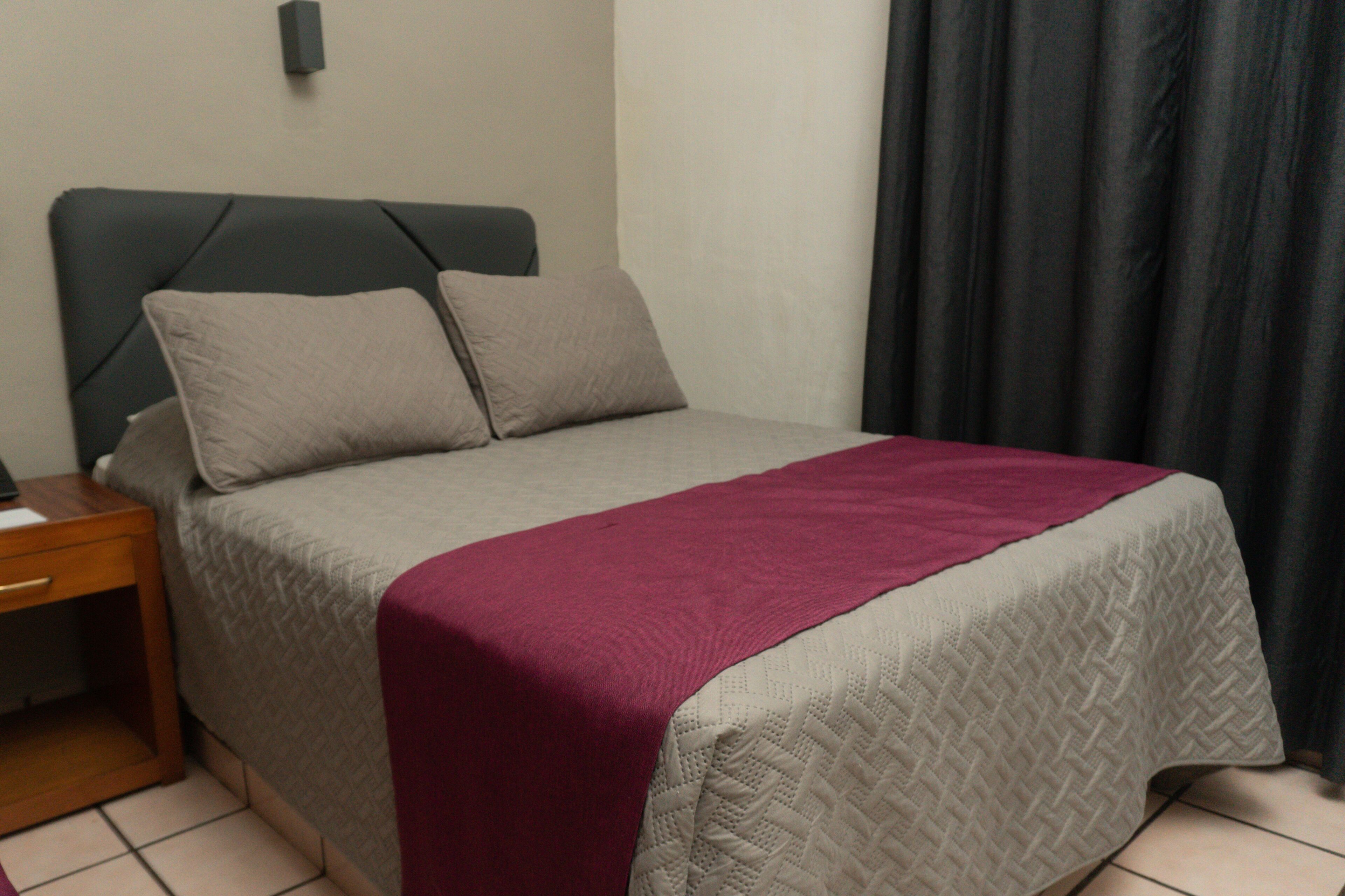 A neatly made bed with a grey and white quilt, a red blanket, and two pillows.