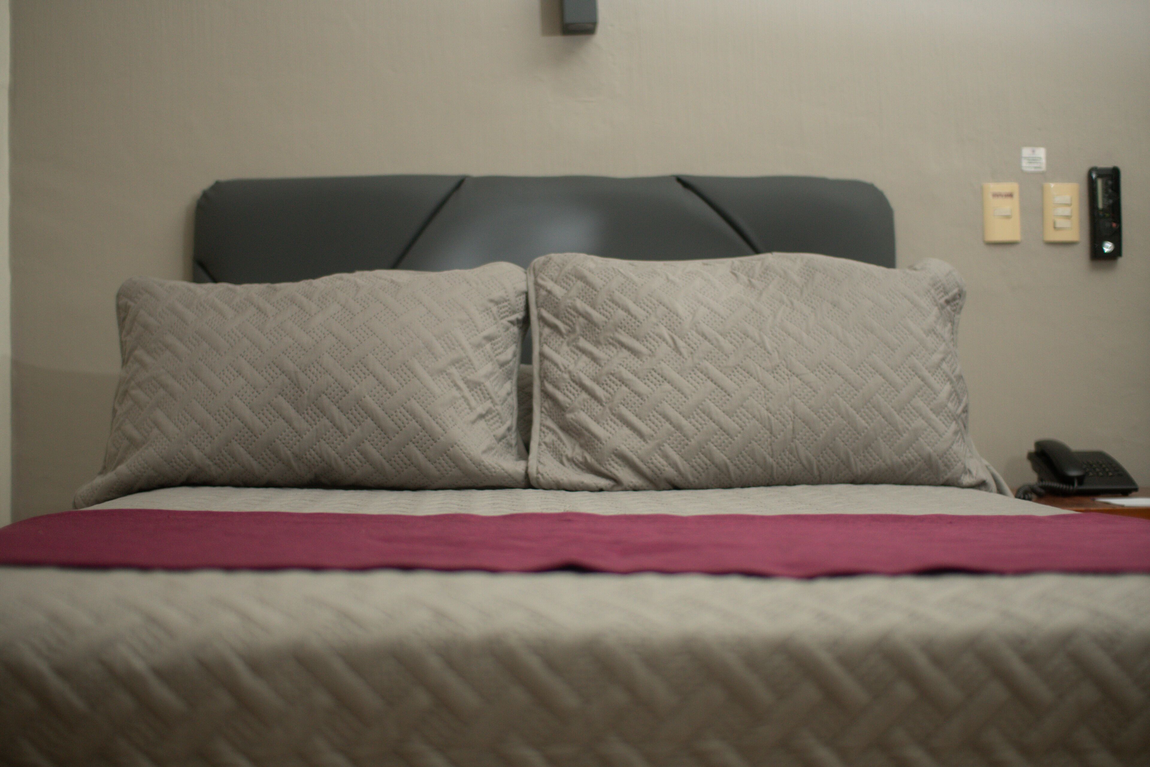 A bed with two pillows, a headboard, and a red blanket. There are two wall-mounted devices on the wall.