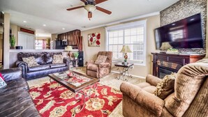 A living room with a ceiling fan, a flat-screen TV, a fireplace, and a large window with blinds. - Little House on the Fairway- Luxury Golf Condo (Branson)