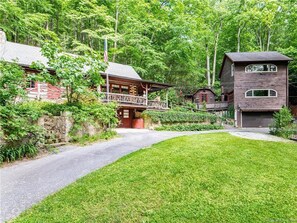 Un chalet rustique doté d’une terrasse en bois, entouré d’une végétation luxuriante et d’un mur de pierres.